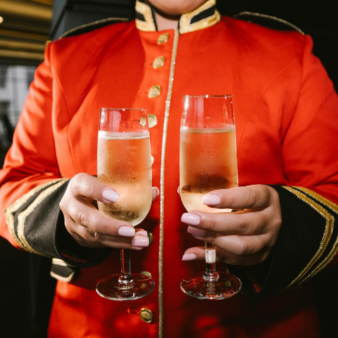 An usher wearing a red coat uniform holds two glasses of champagne in their hands. Their head is cut off.